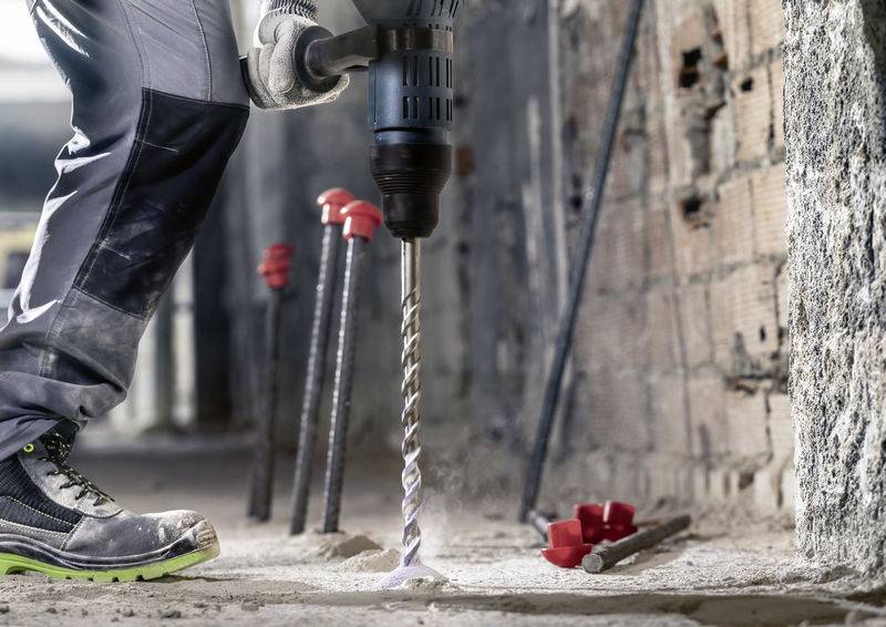 Eine Person bohrt mit einer Bohrmaschine in eine Betonwand auf einer Baustelle. Rote Stifte sind als Markierung in der Wand angebracht.