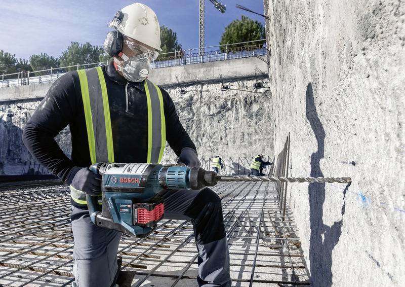 Ein Bauarbeiter trägt Sicherheitsausrüstung und bohrt mit einer elektrischen Bohrmaschine Löcher in eine Betonwand auf einer Baustelle.
