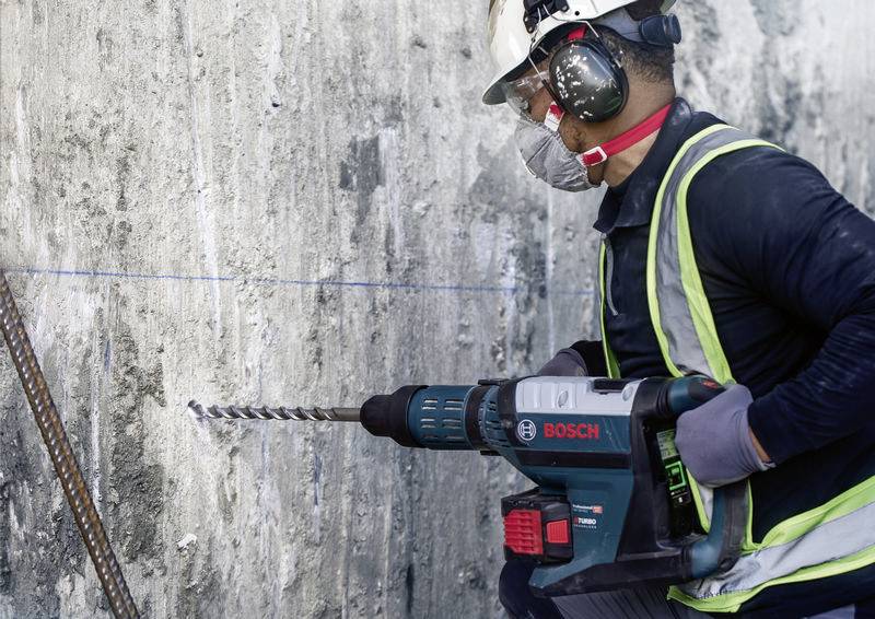 Eine Person in Sicherheitskleidung bedient eine Bosch-Bohrmaschine, um in eine Betonwand zu bohren. Schutzhelm, Gehör- und Augenschutz sind sichtbar.