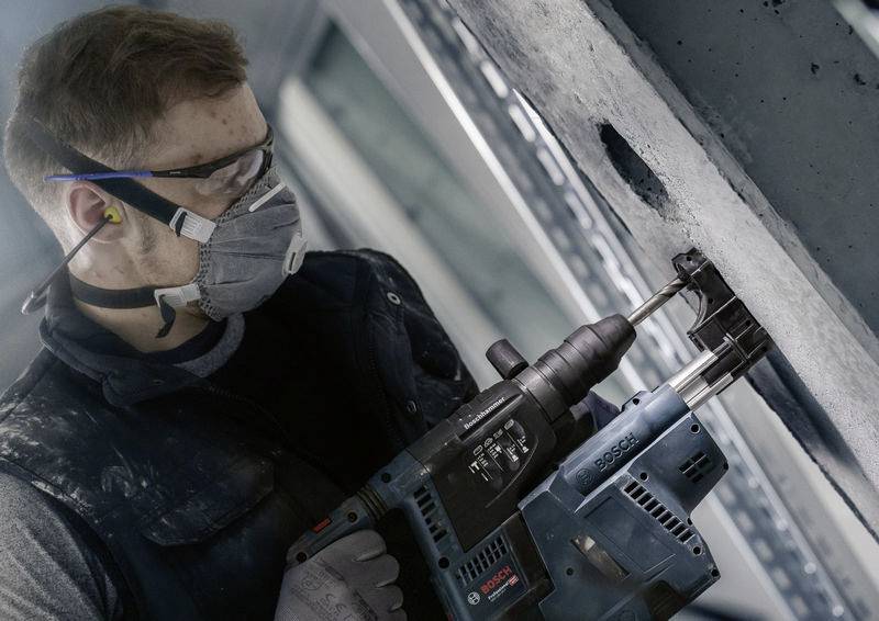 Ein Mann trägt Schutzbrille und Atemschutzmaske, während er mit einem Bohrhammer ein Loch in eine Betonwand bohrt.