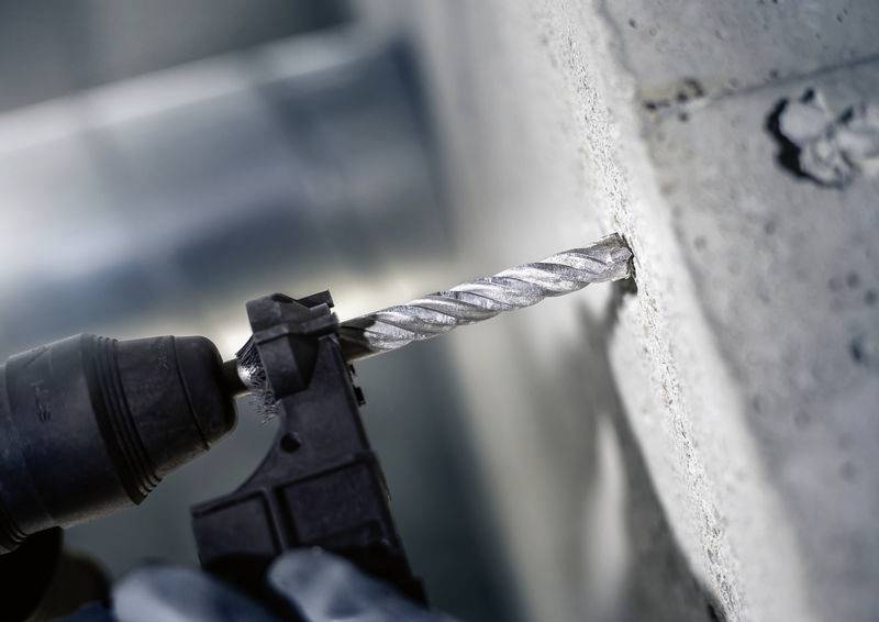 Bohrmaschine mit Metallbohrer bohrt in Betonwand. Nahaufnahme zeigt Bohrer im Einsatz. Arbeitsplatzumgebung sichtbar.