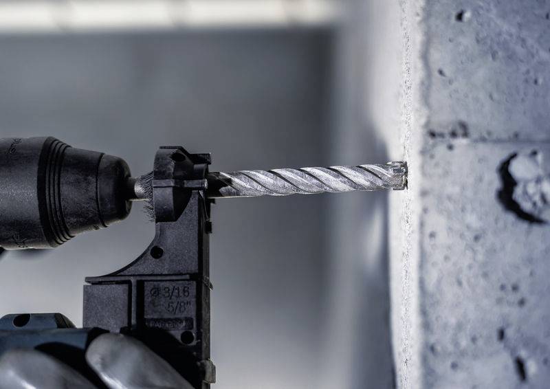 Hand hält Bohrmaschine, die in eine Betonwand bohrt. Fokus liegt auf dem Bohrer, der Staub erzeugt. Hintergrund unscharf.
