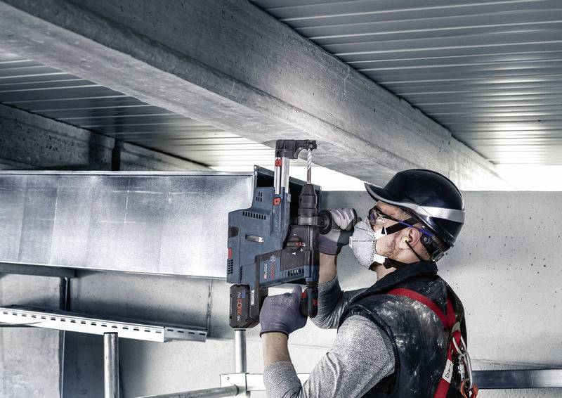 Ein Bauarbeiter mit Schutzhelm und Maske befestigt Metallgestänge unter einer Decke in einem modernen Gebäude.