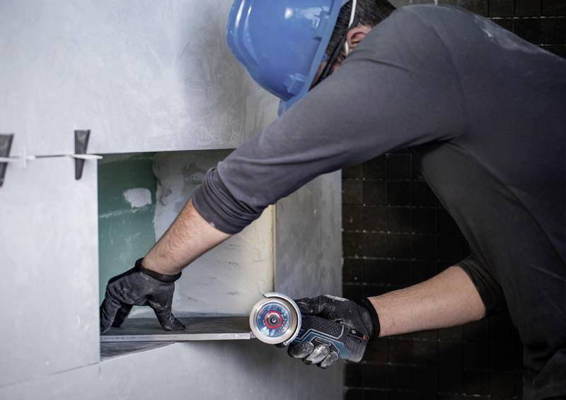 Eine Person trägt einen blauen Sicherheitshelm und schwarze Handschuhe. Sie verwendet ein Elektrowerkzeug, um ein Loch in einer Wand zu vergrößern.