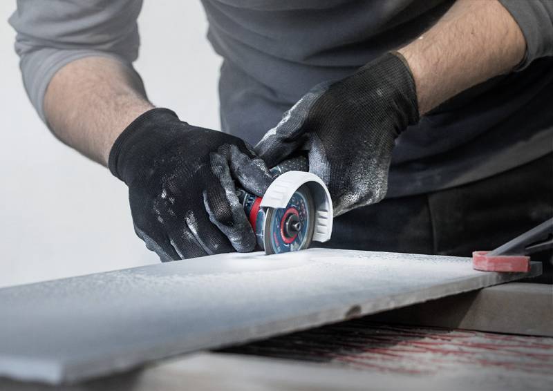 Eine Person arbeitet mit Handschuhen an einer Schleifmaschine, um eine harte Platte zu bearbeiten. Es entsteht Staub durch den Schleifvorgang.