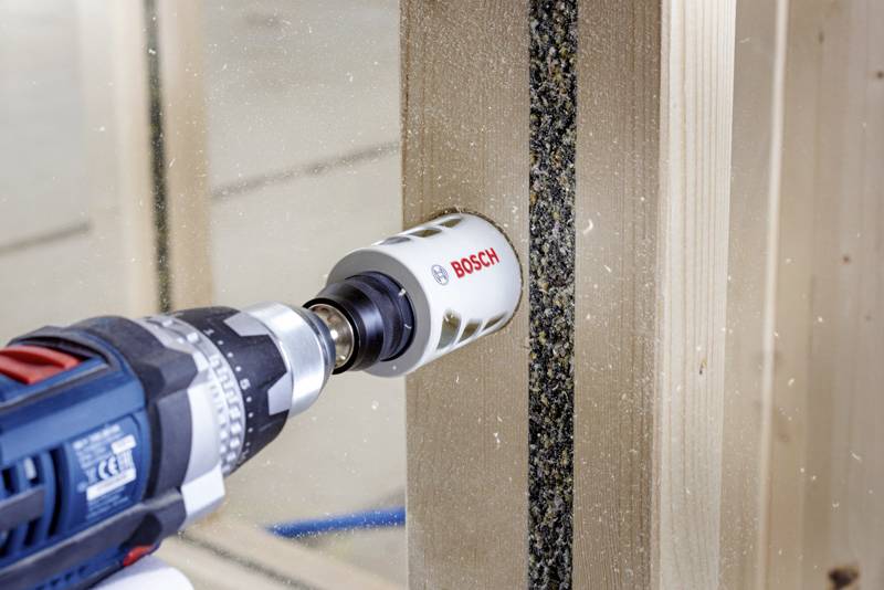 Eine elektrische Bohrmaschine mit einem Lochsägeaufsatz von Bosch bohrt durch einen Holzrahmen und erzeugt Holzspäne.