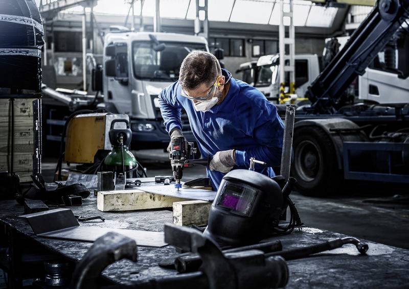 Ein Mensch in blauer Arbeitskleidung und Schutzbrille bohrt auf einer Werkbank in einer Fabrikhalle. Im Hintergrund steht schwere Maschinerie.