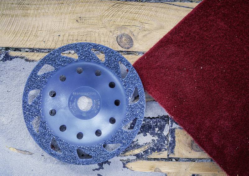 Ein blaues Schleifwerkzeug liegt auf einer Holzbodenfläche, neben einem roten Teppich.