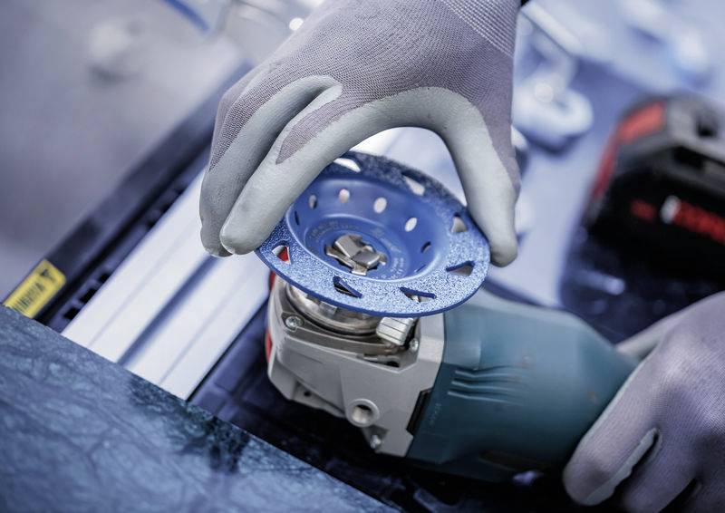 Eine Person mit grauen Handschuhen bedient einen Winkelschleifer mit einer blauen Schleifscheibe. Das Werkzeug wird auf eine Oberfläche gehalten.