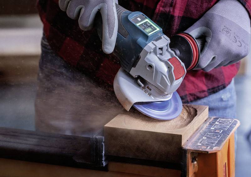 Eine Person schleift mit einer elektrischen Schleifmaschine Holz, wobei Holzspäne in die Luft fliegen. Sie trägt Arbeitshandschuhe.