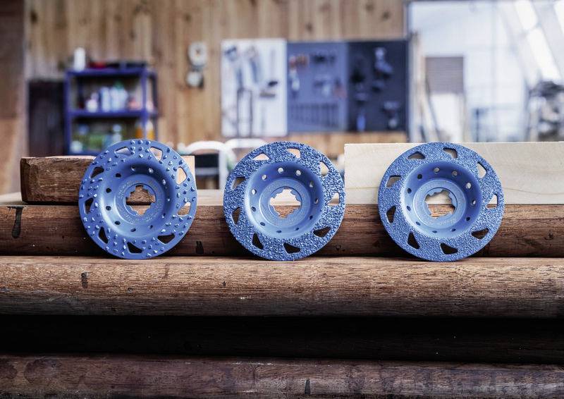 Drei blaue Schleifscheiben mit Löchern liegen nebeneinander auf Stapeln von Holzbalken in einer Werkstatt. Im Hintergrund sind Werkzeuge sichtbar.