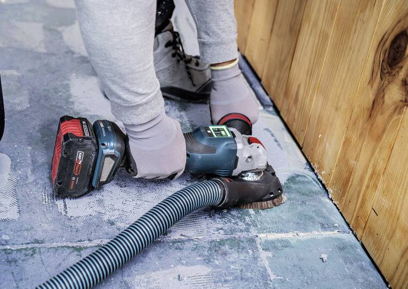 Person benutzt elektrische Schleifmaschine auf einem Boden neben einer Holzwand. Staubabsaugschlauch ist angeschlossen.