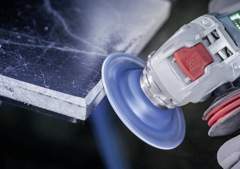 Eine Schleifmaschine bearbeitet die Kante eines Metallstücks. Die rotierende blaue Schleifscheibe erzeugt Funken und Glanz an der Kante.