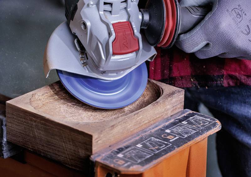 Eine Person schleift mit einem Winkelschleifer eine Holzoberfläche. Das Werkzeug dreht sich schnell und sprüht leichte Späne ab.