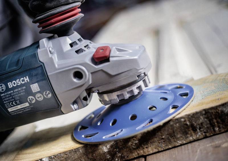 Eine elektrische Schleifmaschine schleift eine Holzoberfläche. Die Schleifscheibe ist blau, die Maschine grau mit rotem Knopf.