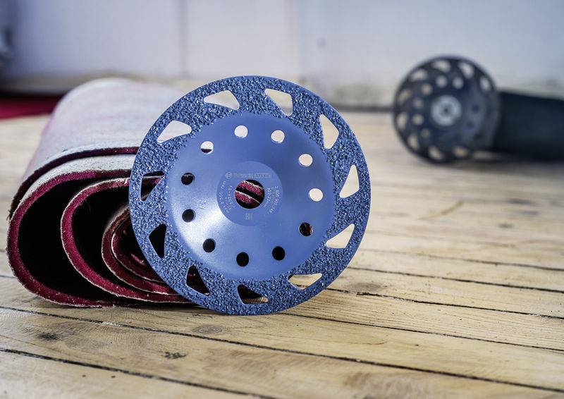 Ein blauer Schleifteller liegt auf einem gerollten Teppich auf einem Holzboden. Im Hintergrund ist ein weiterer Schleifteller sichtbar.
