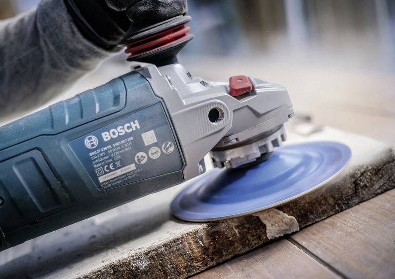 Eine Person schleift eine Holzoberfläche mit einem Winkelschleifer der Marke Bosch, der blauen Schleifscheibenaufsatz verwendet.