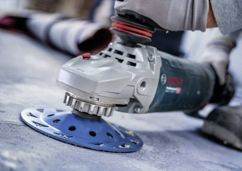 Eine Hand hält eine elektrische Schleifmaschine mit einer blauen Schleifscheibe. Sie schleift eine Betonoberfläche.