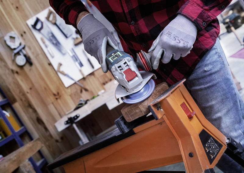 Person schleift ein Holzstück mit einem Winkelschleifer, trägt Handschuhe und ein kariertes Hemd. Werkstatt mit Werkzeug im Hintergrund.