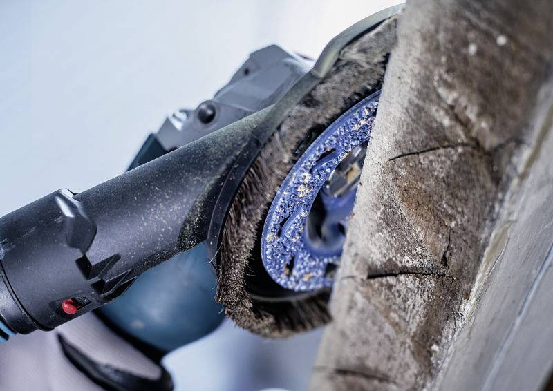 Eine blaue elektrische Schleifmaschine bearbeitet eine abgenutzte Holzkante. Die rotierende Scheibe entfernt alte Lack- oder Schmutzschichten.