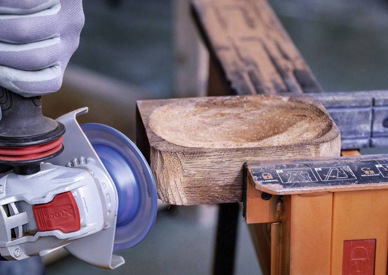 Person bearbeitet ein Holzstück mit einer Schleifmaschine. Das Holz ist auf einer Werkbank befestigt. Schutzhandschuhe werden getragen.