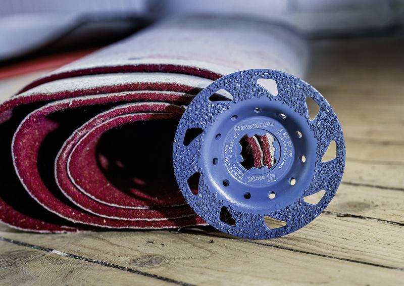 Ein zusammengerollter Teppich liegt auf einem Holzboden. Daneben befindet sich eine blaue Schleifscheibe, die für Bodenarbeiten verwendet wird.