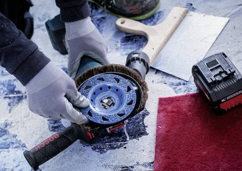Eine Person mit Handschuhen hält ein Schleifwerkzeug und bereitet es auf eine Arbeitsfläche vor. Neben dem Werkzeug liegt eine Batterie.