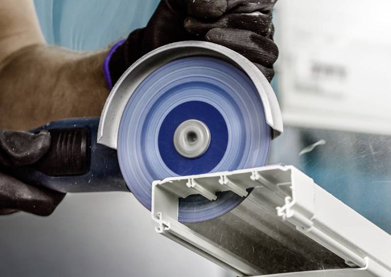 Person schneidet mit einer blauen Kreissäge ein Metallstück. Schutzbekleidung ist sichtbar, Konzentration auf Präzision beim Schneiden.