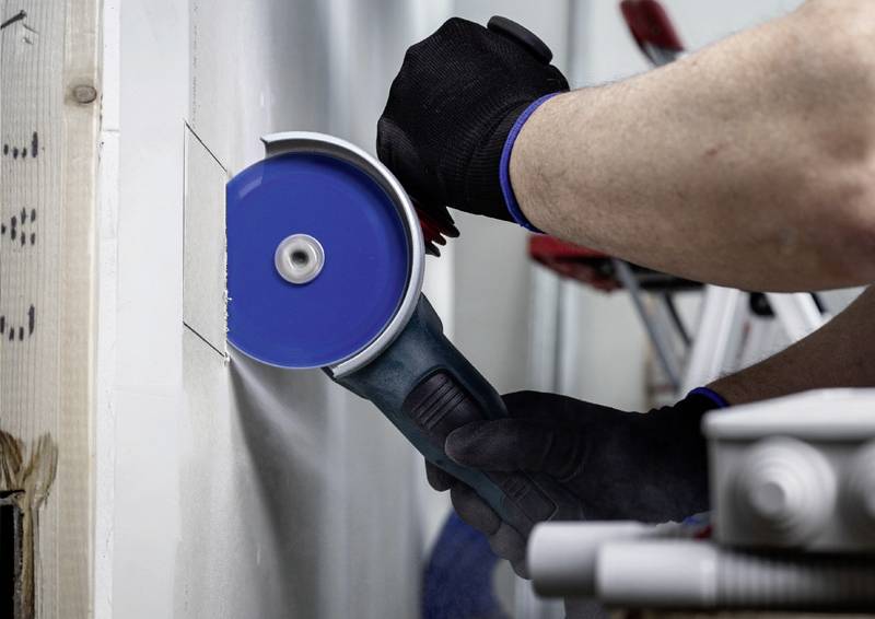 Eine Person schneidet mit einer blauen Kreissäge ein Loch in eine weiße Wand. Sie trägt schwarze Handschuhe.