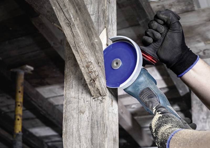 Handwerker benutzt Winkelschleifer, um einen Holzbalken zu bearbeiten. Trägt Schutzhandschuhe und arbeitet in einer Holzkonstruktion.