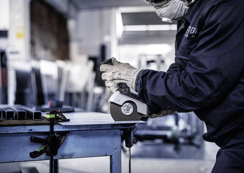 Eine Person in Schutzkleidung schleift Metall auf einem Werktisch. Im Hintergrund unscharfe Werkstattumgebung.