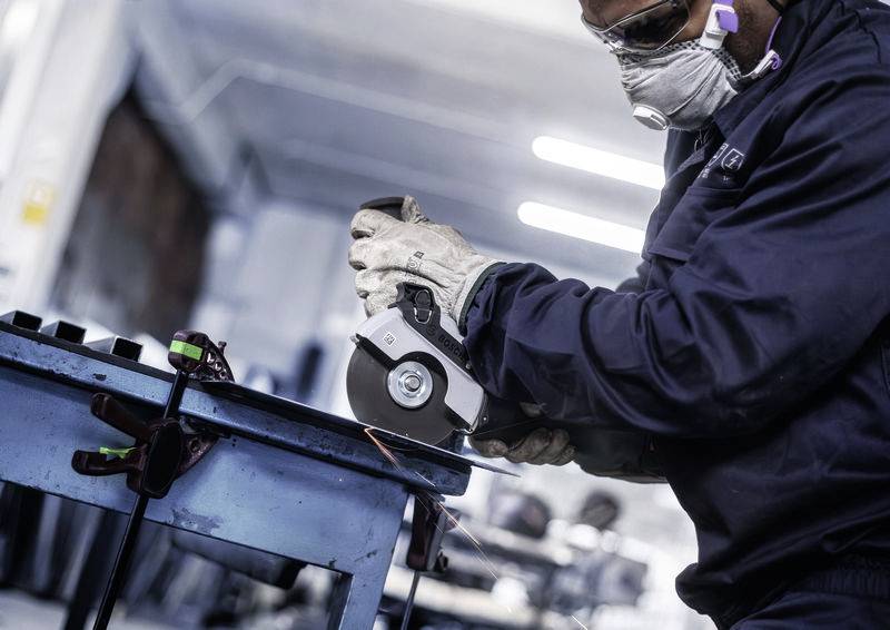 Arbeiter mit Schutzbrille und Maske schleift Metall in einer Werkstatt. Starkes Licht im Hintergrund.