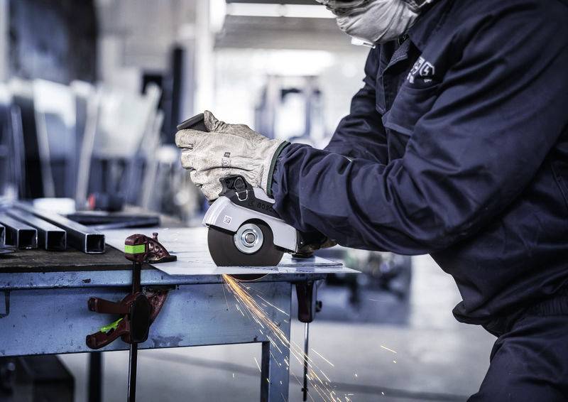 Eine Person in Arbeitskleidung schneidet mit einem Winkelschleifer Metall auf einem Tisch. Funken sprühen, während sie arbeitet.