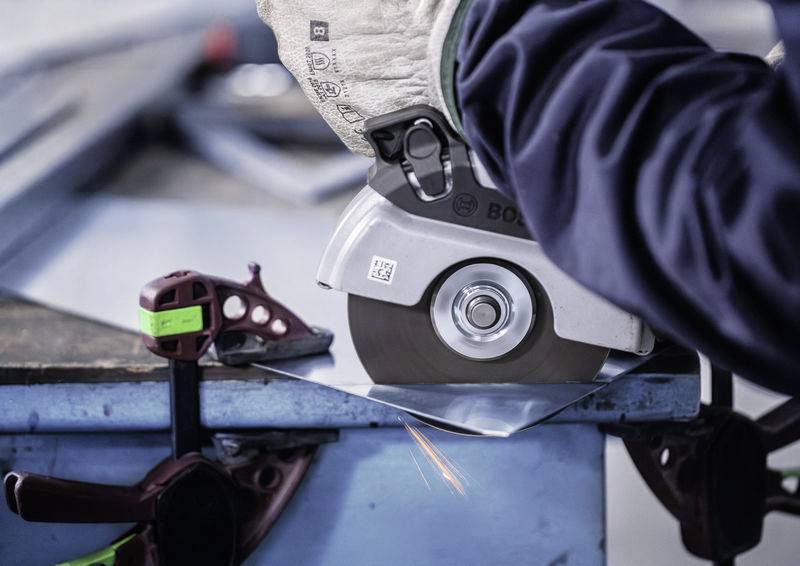 Eine elektrische Schleifmaschine bearbeitet eine Metallplatte auf einem Tisch, Funken sprühen. Eine Klemme fixiert das Material.