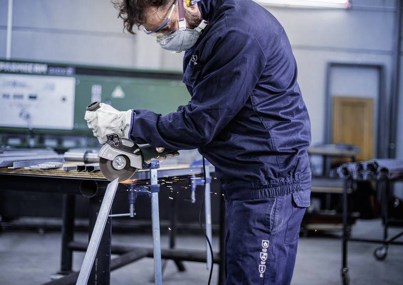 Eine Person in Schutzkleidung schleift mit einem Winkelschleifer Metall in einer Werkstatt. Funken fliegen vom Metall weg.