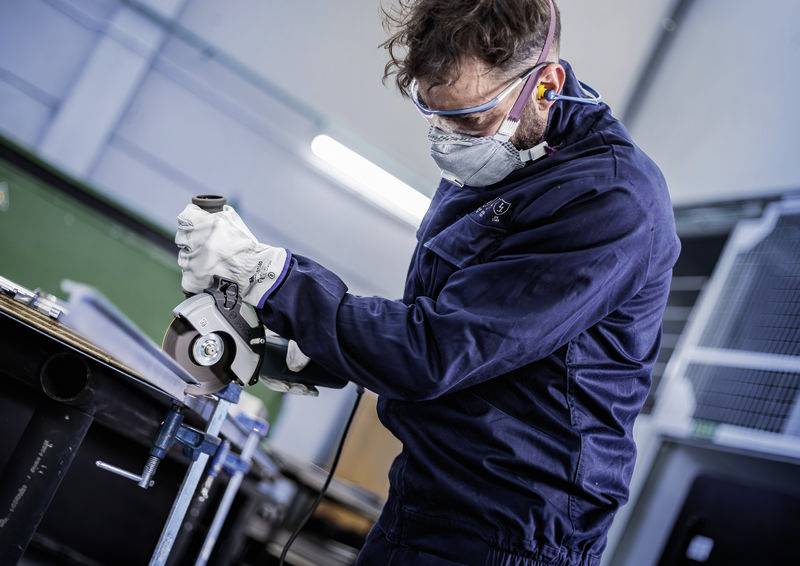 Eine Person in Schutzkleidung schleift Metall mit einem Winkelschleifer in einem Industriebetrieb.