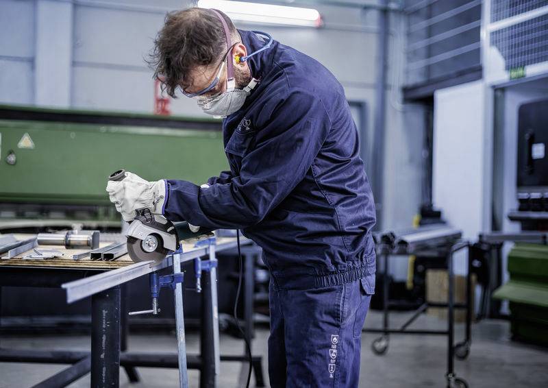 Ein Mann in Arbeitskleidung schleift Metall in einer Werkstatt. Er trägt Schutzbrille, Gehörschutz und eine Maske für Sicherheit.