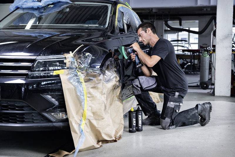 Ein Mann kniet und poliert mit einem Gerät das linke Vorderteil eines schwarzen Autos; Reparatur- oder Pflegesituation in der Werkstatt.