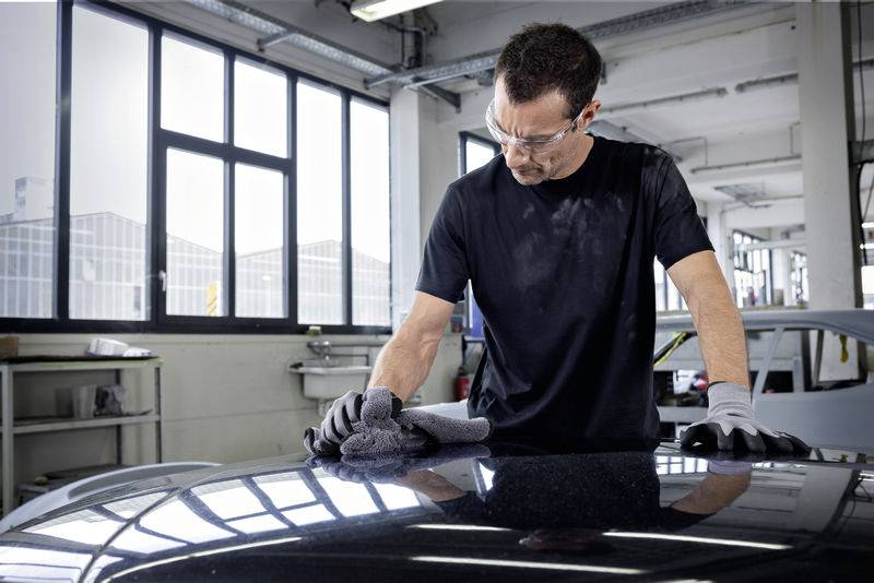 Ein Mann in einer Werkstatt poliert ein Auto mit einem Tuch, trägt Schutzbrille und Handschuhe.