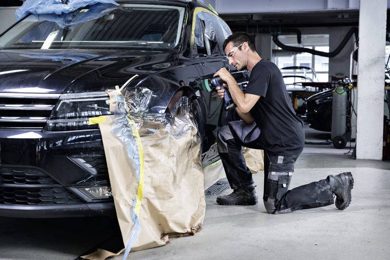 Ein Mann kniet neben einem schwarzen Auto und setzt mit einem Werkzeug die Karosserie instand, um den Lack auszubessern oder zu reparieren.