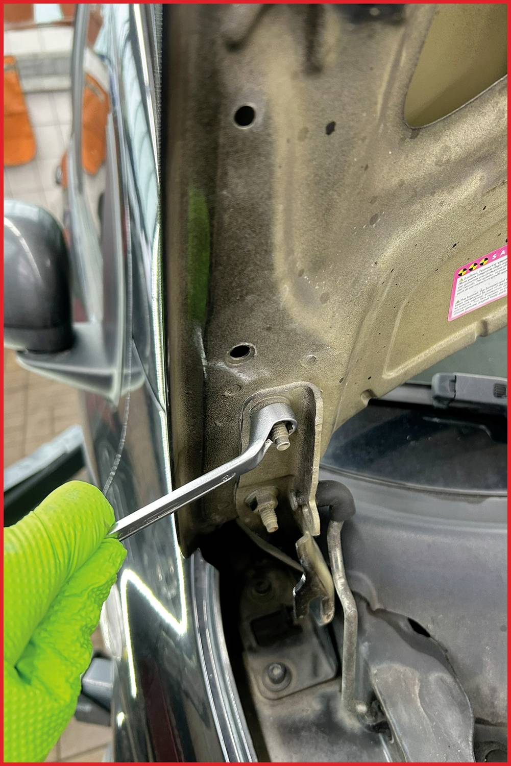 Grüne behandschuhte Hand schraubt mit einem Inbusschlüssel eine Schraube an einem Autotürgelenk fest.