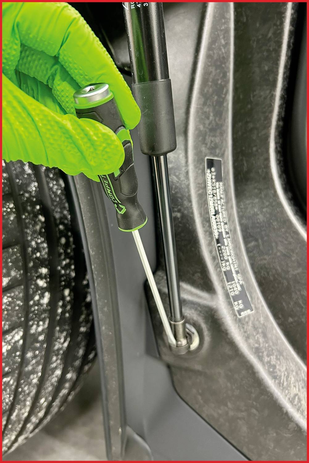 Hand in einem grünen Handschuh, das einen Schraubendreher hält und eine Schraube an einem Metallteil in einem Fahrzeug befestigt.