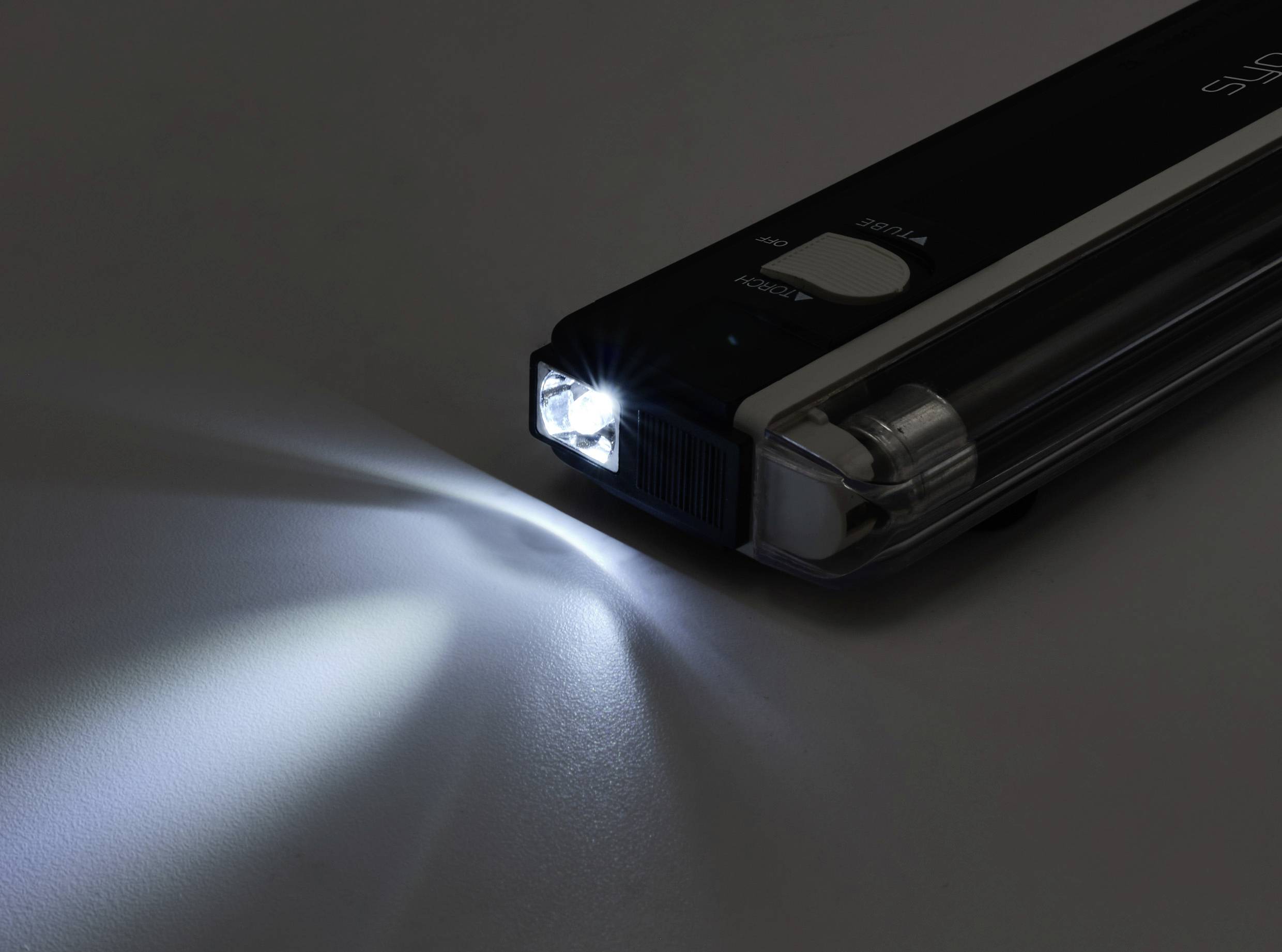 Eine kompakte Taschenlampe, die auf einer Oberfläche liegt und einen hellen Lichtstrahl nach links aussendet, wobei ein Teil der Umgebung beleuchtet wird.