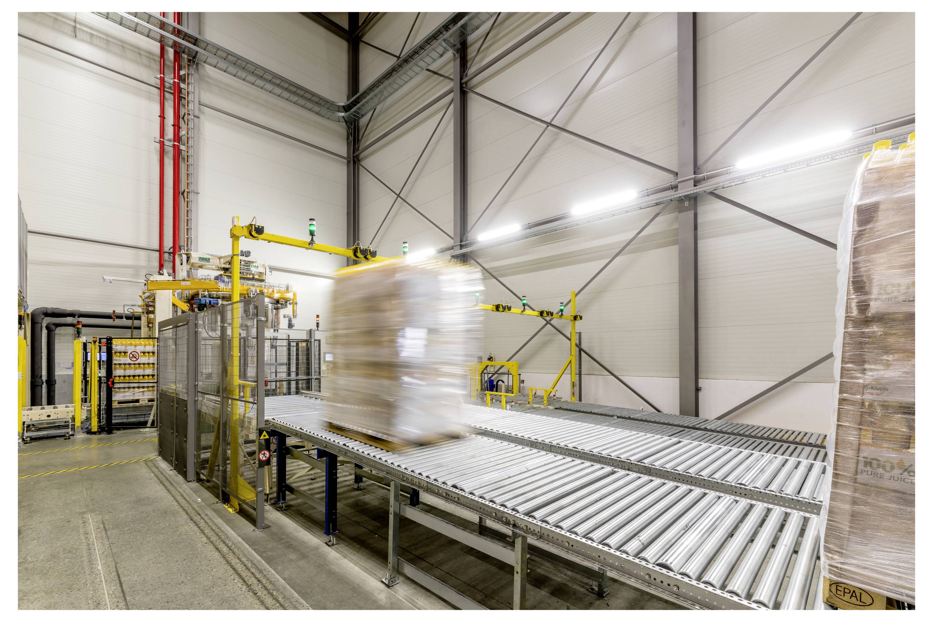 Eine Lagerfoto zeigt eine umwickelte Palette auf einem Transportsystem, was automatisierte Warenlogistik in einem industriellen Umfeld andeutet.