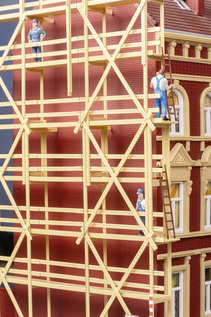 Drei Arbeiter auf einem Baugerüst an einem Backsteingebäude. Einer steht oben, die anderen sind weiter unten. Sie tragen Schutzhelme.