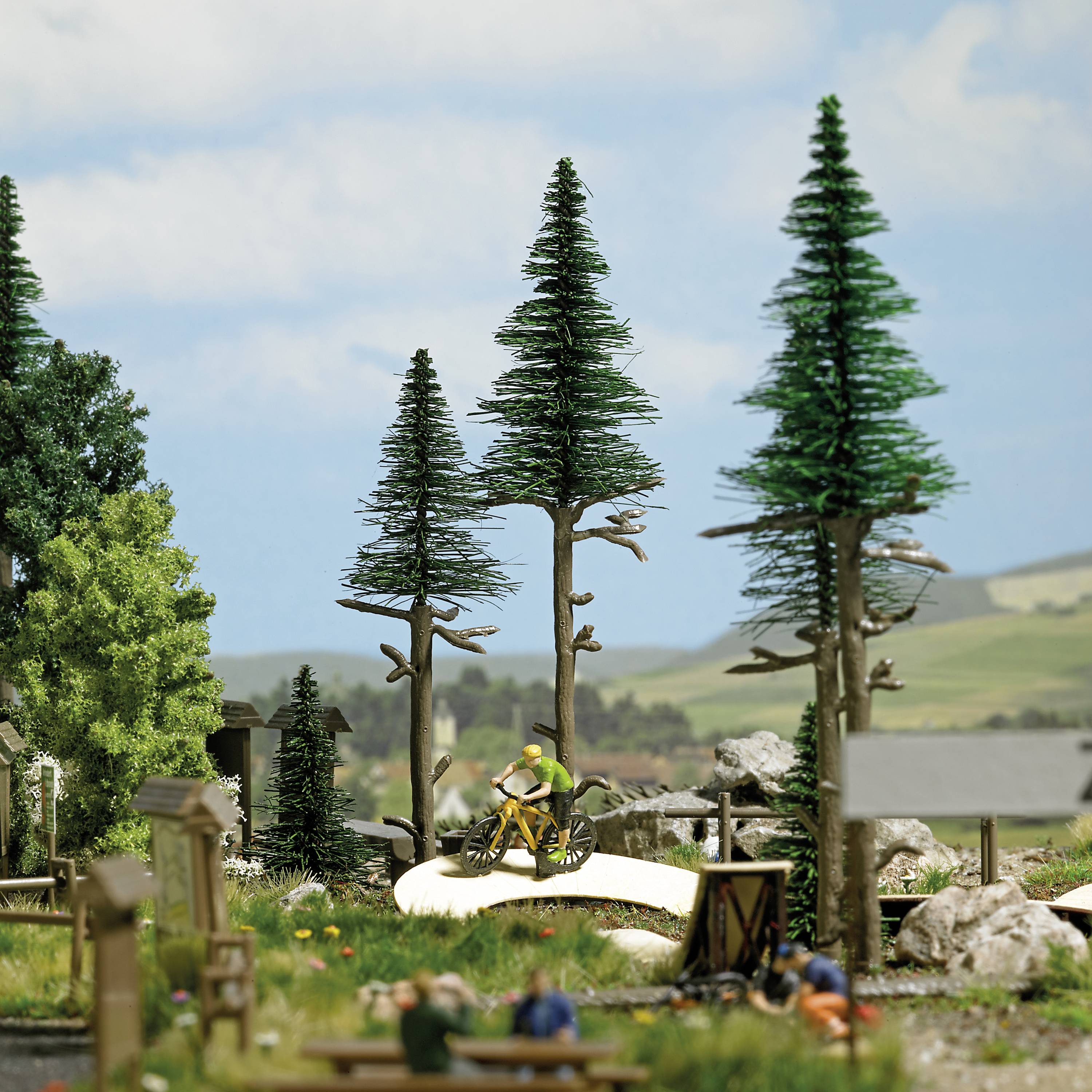 Eine Miniaturlandschaft zeigt einen kleinen Park mit hohen Bäumen und zwei Radfahrern auf einem gewundenen Weg. Im Hintergrund sind Hügel sichtbar.
