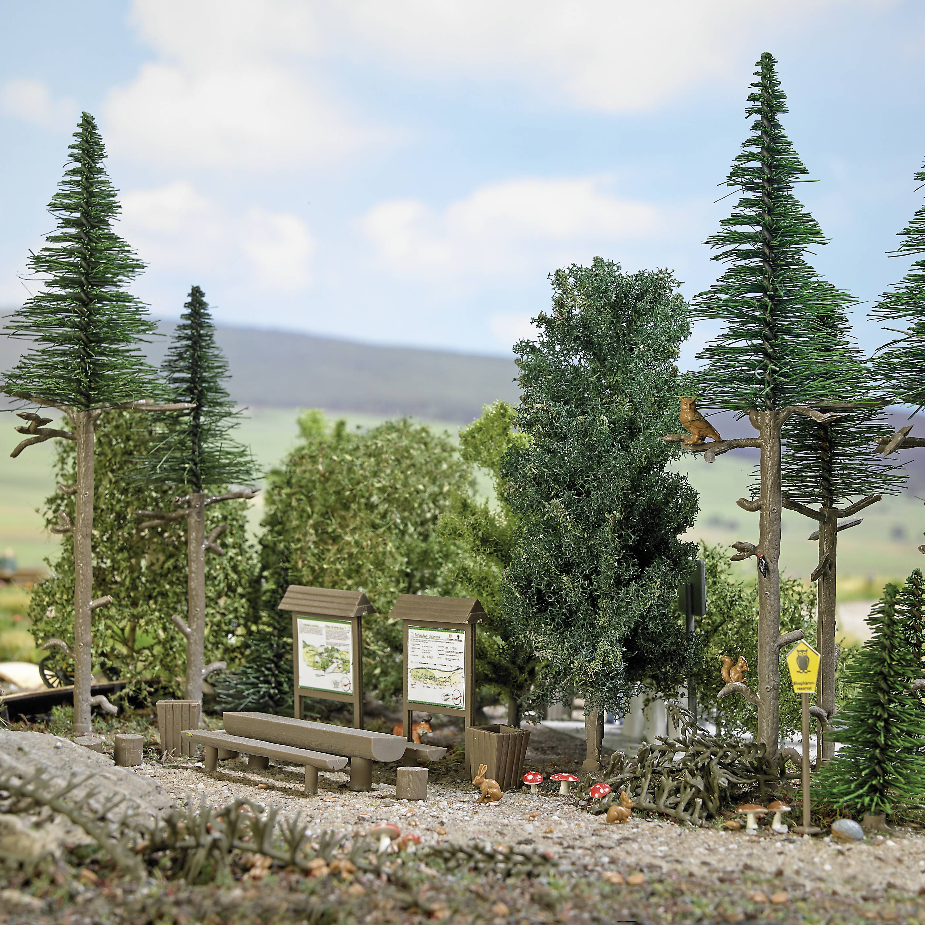 Ein Miniatur-Waldabschnitt mit hohen Bäumen, einem Picknicktisch und Informationstafeln. Szenerie wirkt entspannend und friedlich.