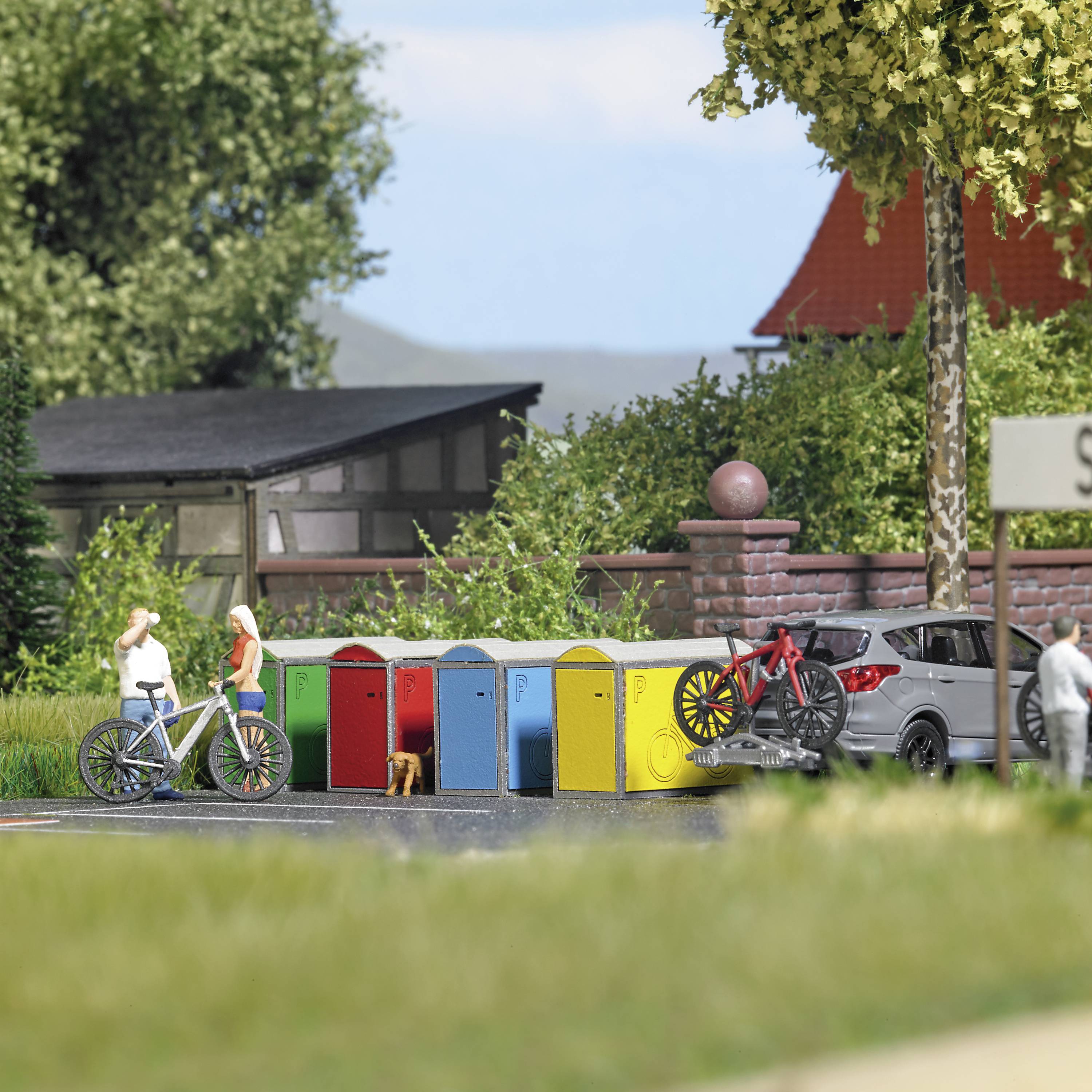 Eine bunte Reihe von Fahrradboxen steht in einer Wohngegend. Zwei Personen mit Fahrrädern unterhalten sich davor.