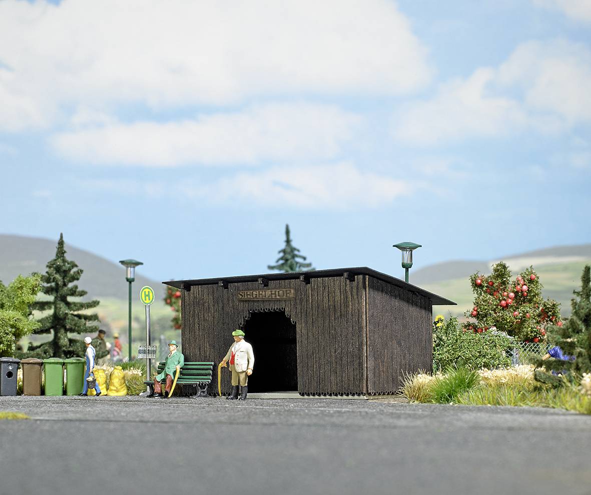 Modell einer Bushaltestelle mit angrenzender Holzhütte, Bänken und Figuren in ländlicher Umgebung mit Bäumen und Hügeln im Hintergrund.