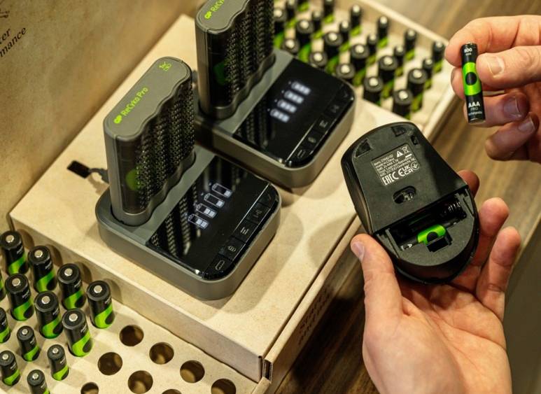 Eine Hand hält ein Batteriefach und legt eine aufladbare Batterie ein, während im Hintergrund ein Ladegerät mit mehreren geladenen Batterien zu sehen ist.